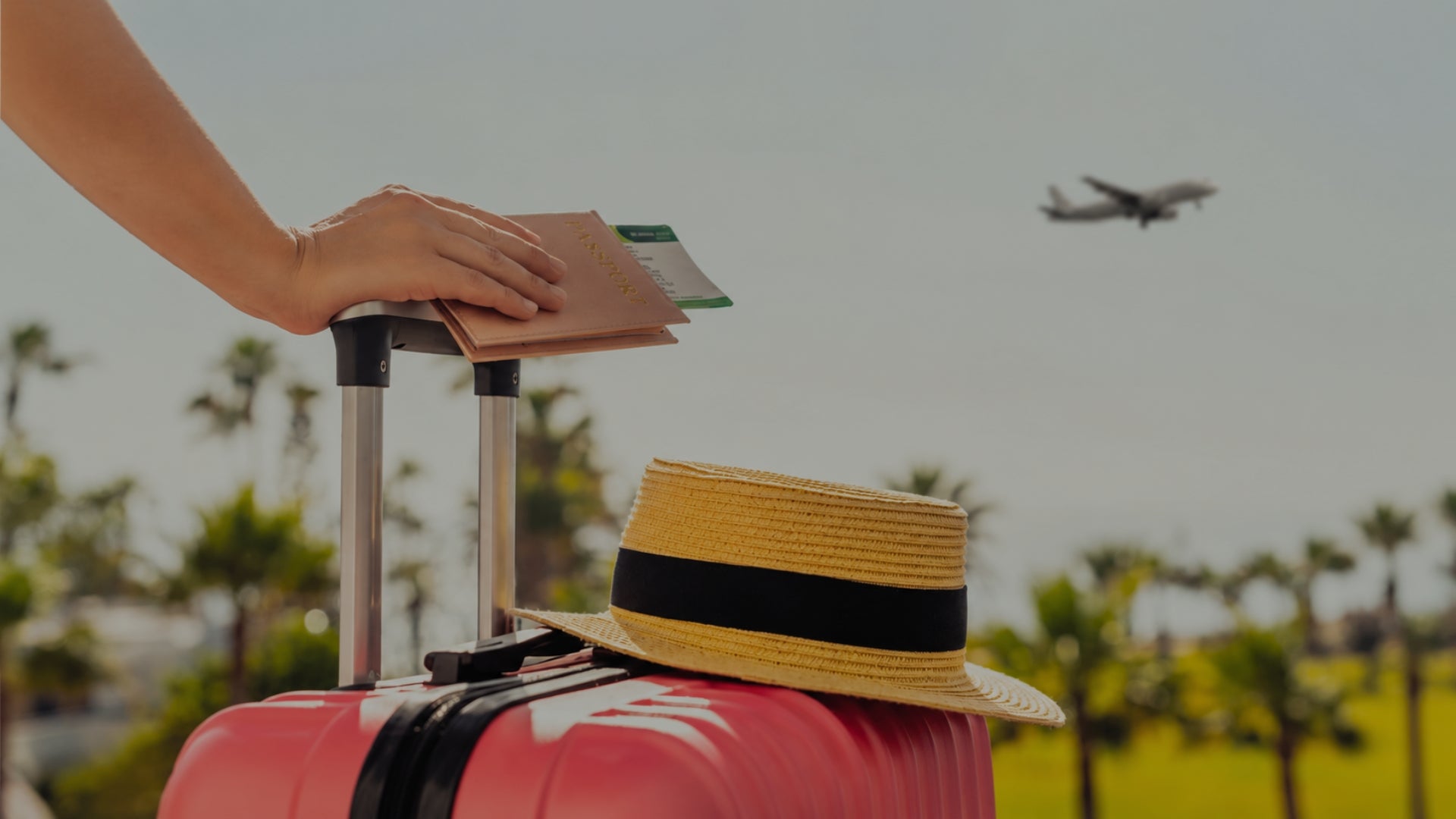 Person holding a passport next to a suitcase with a straw hat on a beach, airplane in the background